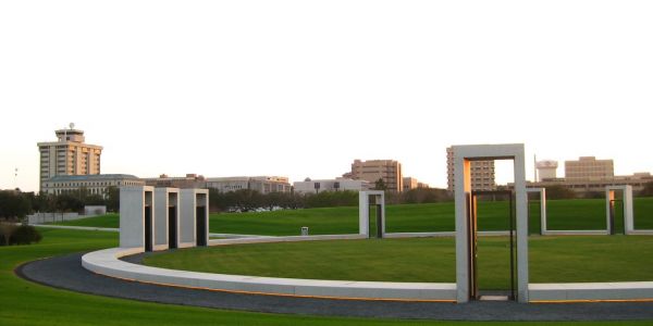 A wide view of a grassy field with modern rectangular sculptures and buildings in the background at sunset near The Arbors at Wolf Pen Creek Apartments.