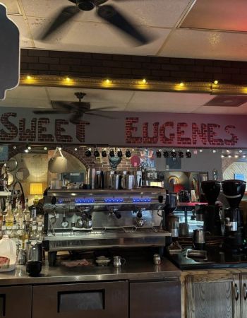 Coffee shop counter with espresso machines, syrup bottles, and a sign reading "SWEET EUGENES" in the mirror near The Arbors at Wolf Pen Creek apartments.